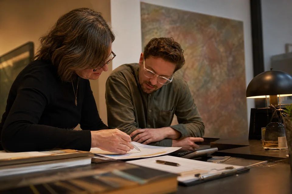 A gallery owner and her associate go through a printed yearly software budget with a calculator at the front desk after closing