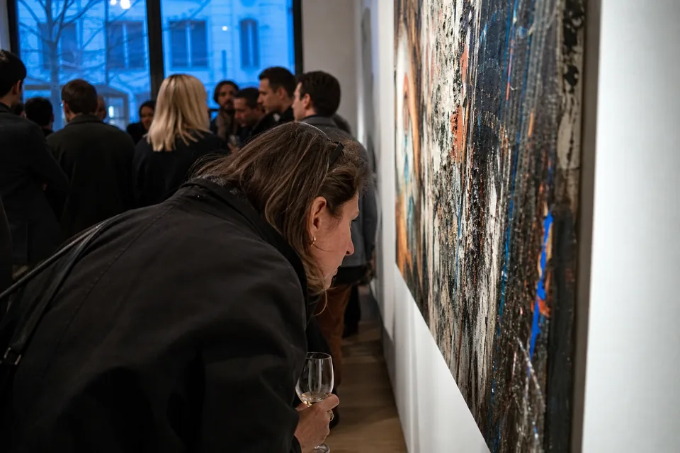 Over the shoulder view of a collector leaning in to look closely at a painting during a crowded opening night