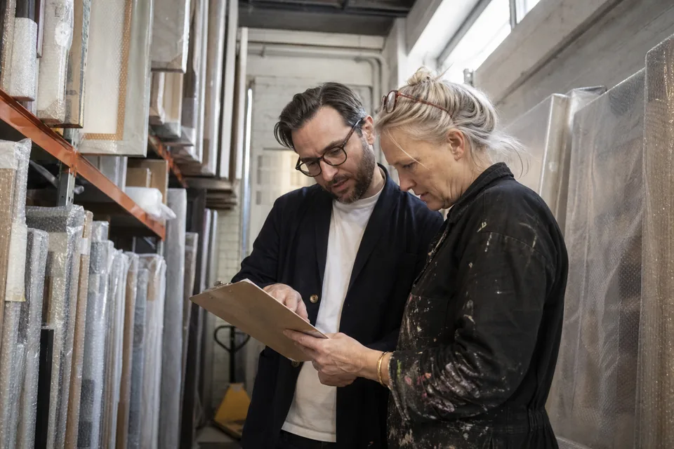 A gallery registrar and an artist check a consignment checklist together beside wrapped canvases in a storage room