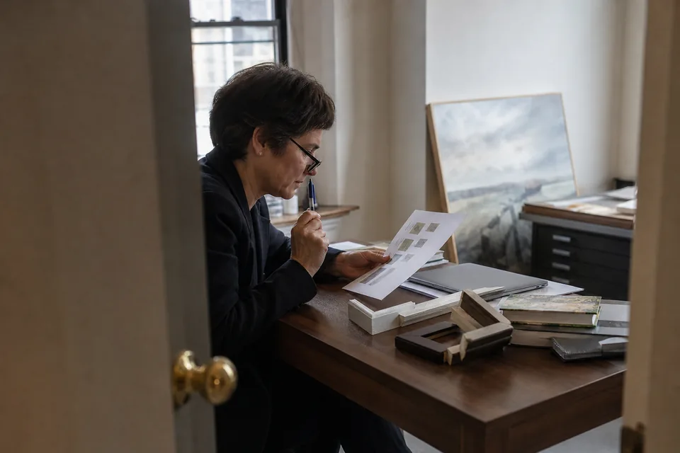 A gallery director at a desk in the back office reads a printed availability list, frame corner samples and a laptop beside her