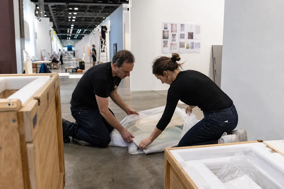 A gallery booth at an art fair during installation, open wooden crates, a printed works list taped to the wall and two staff unwrapping a painting