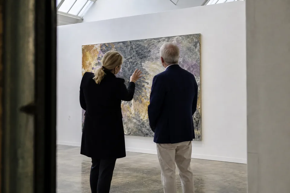 A gallerist walks a collector through a new exhibition, both looking at a large painting on the wall