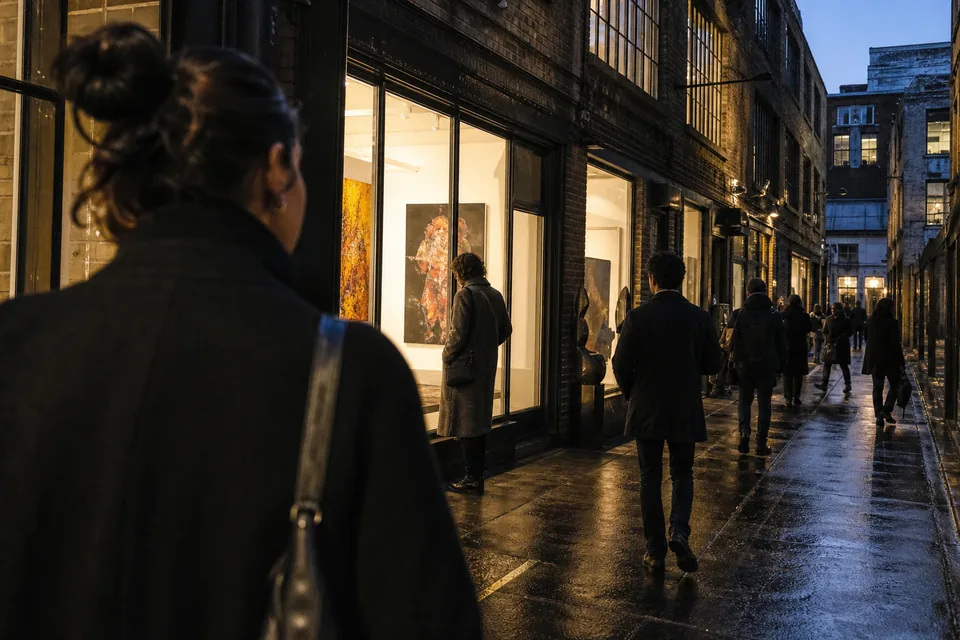 A street in a gallery district at dusk with lit gallery windows showing paintings, people walking between the doors