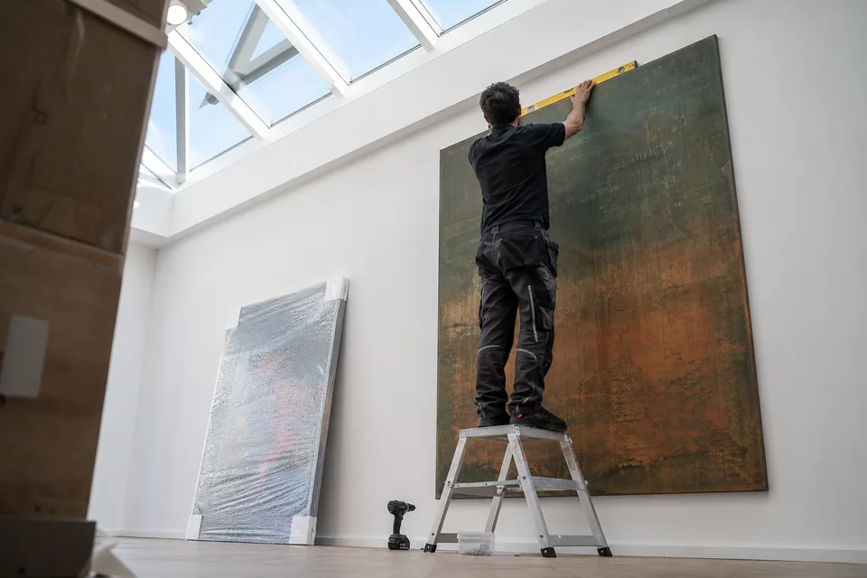 An art handler on a short ladder levels a large abstract painting on a white gallery wall, spirit level in hand
