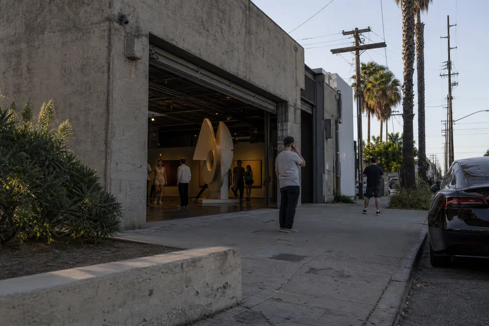 A low warehouse gallery with a roll up door open to the street and palm trees in late afternoon light