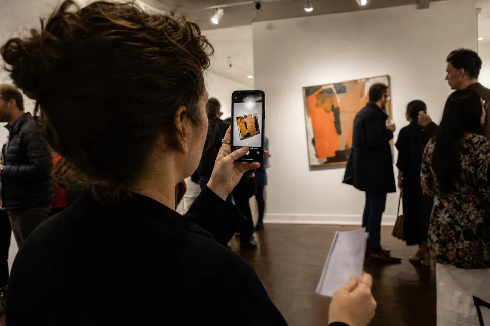 A gallery assistant photographs a painting with her phone during an opening, the crooked snapshot visible on the screen, a folded note in her other hand