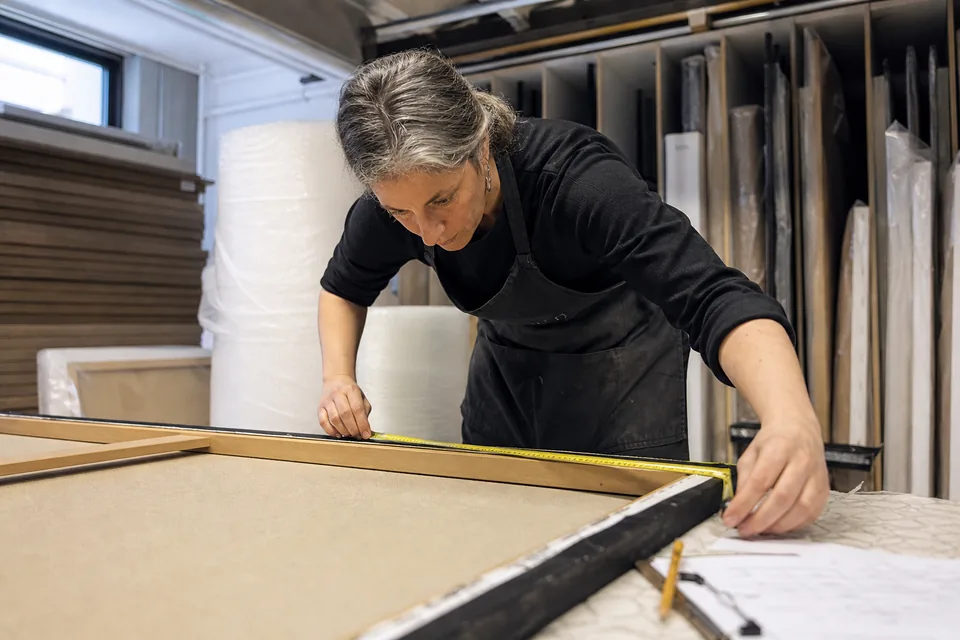 A gallery registrar measures a stretched canvas with a tape measure on a work table in the art storage room, cardboard and bubble wrap behind her