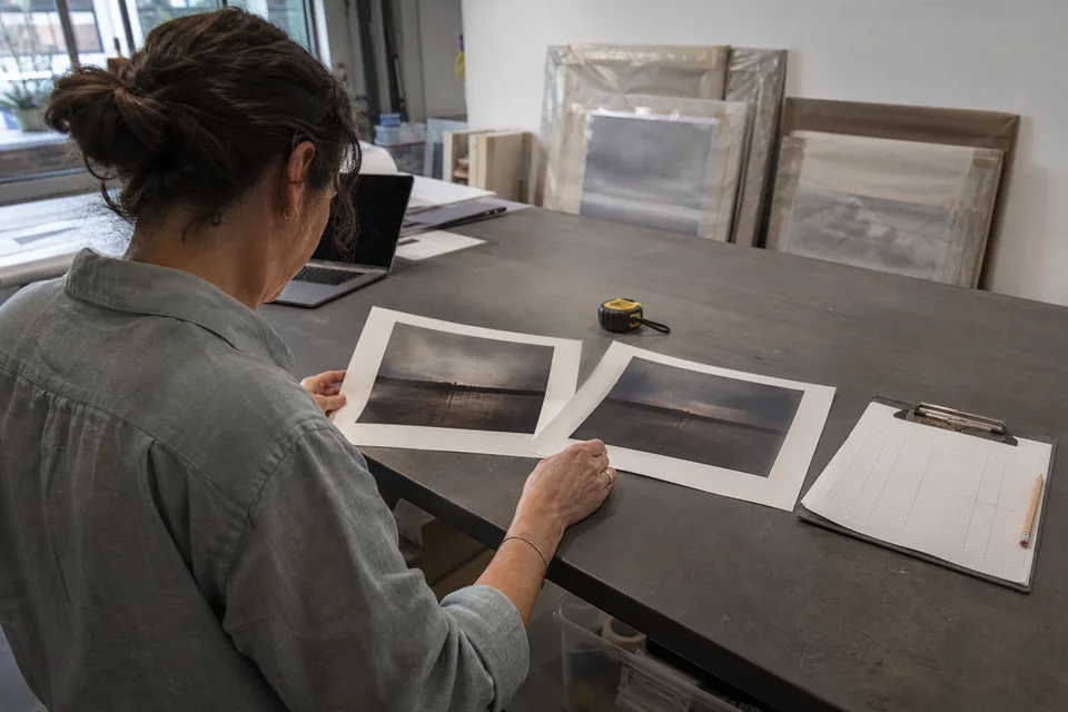 A gallery registrar compares two printed inventory reports at a back office table with a laptop and framed works stacked against the wall